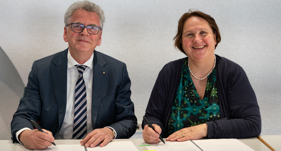 Ein Mann und eine Frau sitzen nebeneinander an einem Tisch, sie halten Stifte in der Hand und vor ihnen liegen zwei Blätter auf dem Tisch. Sie schauen in die Kamera. 