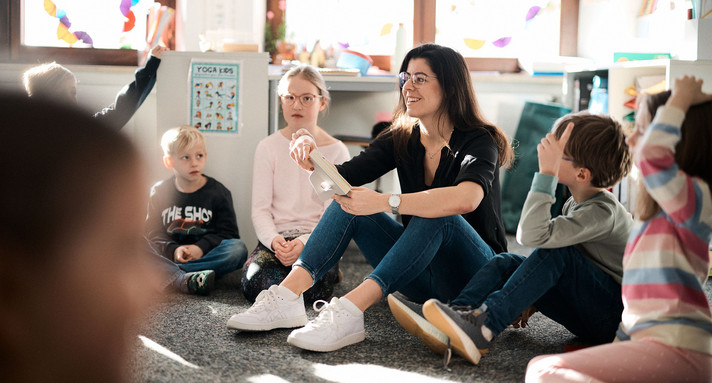 Das Bild zeigt eine Erzieherin, die umgeben von mehreren Kindern, die sich melden, auf dem Boden sitzt.