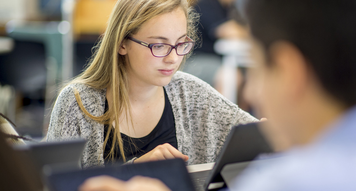 Junge Frau mit Brille mit Tablet im Focus im Klassenraum mit anderen Schülern