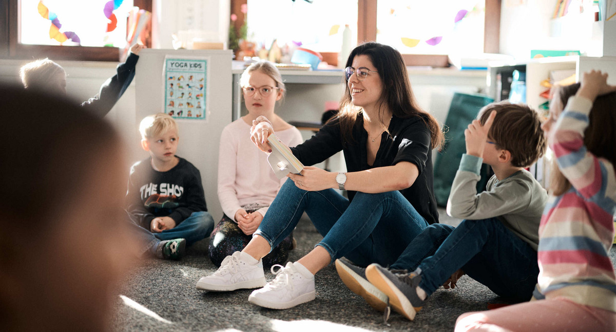 Schülerinnen und Schüler sitzen mit einer Lehrerin in einem Klassenzimmer im Kreis auf dem Boden.