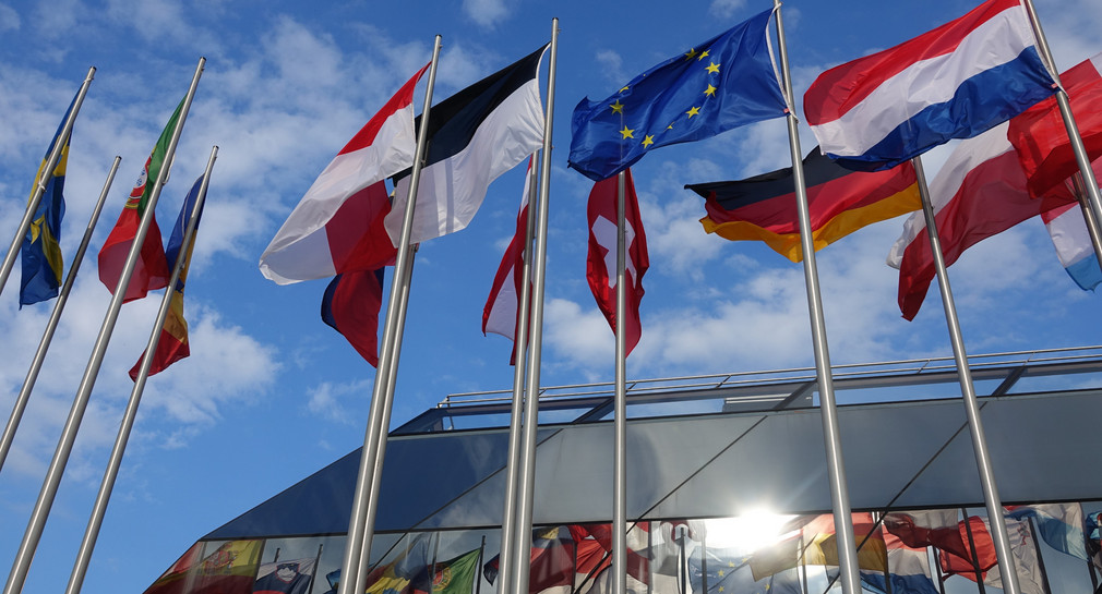Flaggen europäischer Länder wehen vor dem Europaparlament und spiegeln sich in Fenstern.