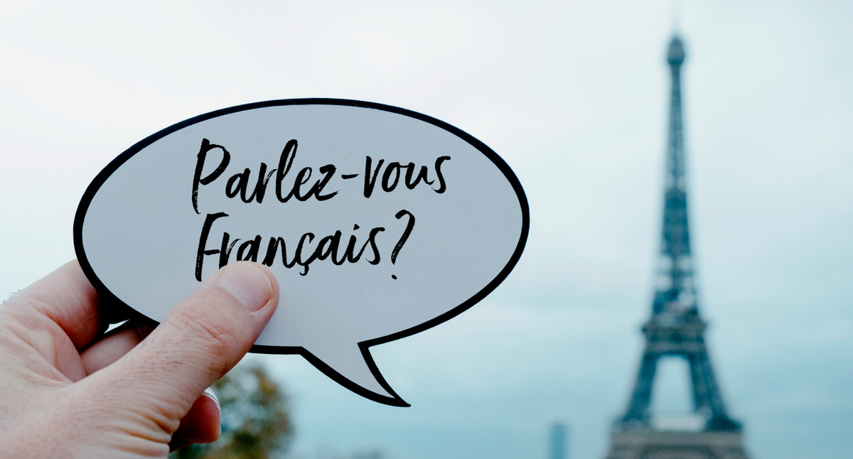 IM Hintergrund sieht man einen Turm aus Eisen, im Vordergrund hält eine Hand ein Schild in die Luft, auf dem Steht "Parlez-vous francais?". 
