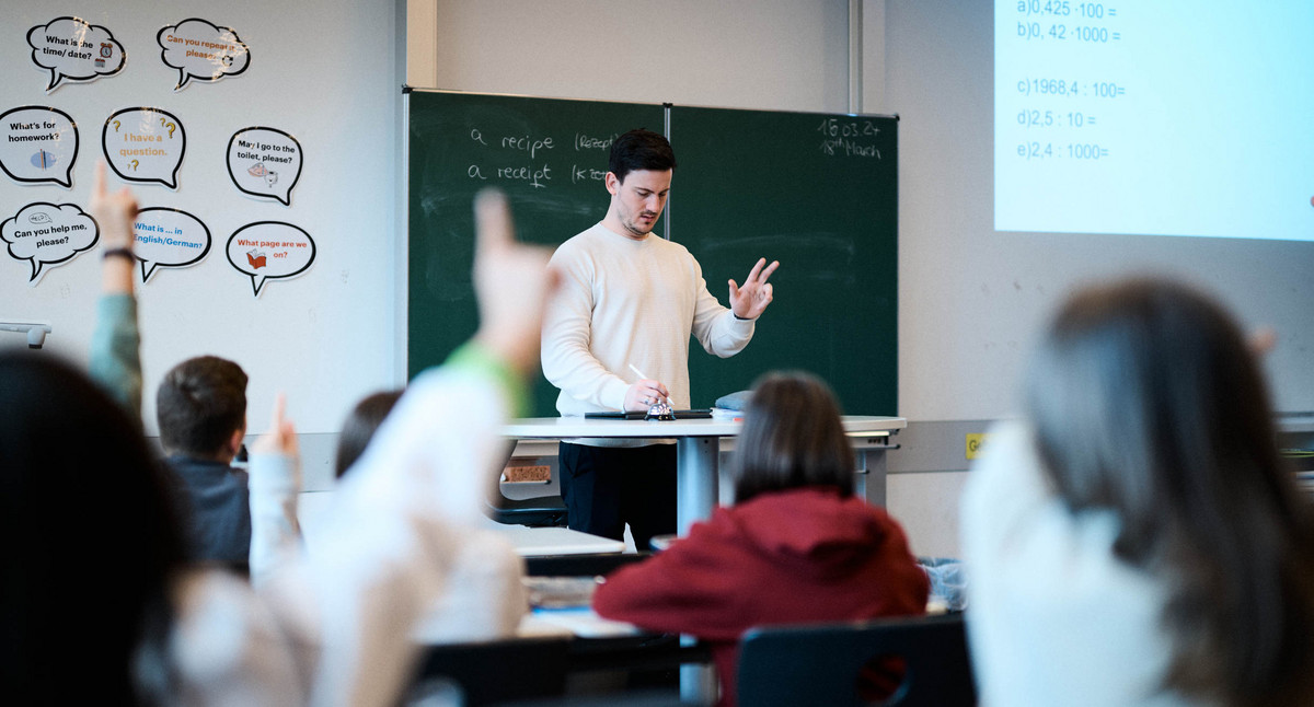 Ein Lehrer steht in einem Klassenzimmer und schreibt auf einem Tablet. Ihm gegenüber sitzen Schülerinnen und Schüler.