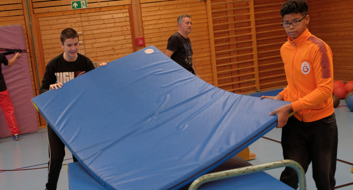 Zwei Schüler heben in der Turnhallte eine blaue Turnmatte auf den Mattenwagen. Im Hintergrund ist ein Lehrer und ein weiterer Schüler zu sehen.