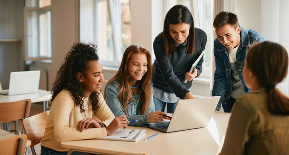 Glückliche Schülergruppe, die mit ihrer Informatiklehrerin während des Unterrichts einen Laptop benutzen