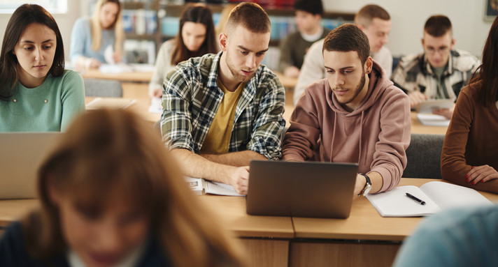 Älterne Gymnasiasten und Gymansiastinnen im Klassenraum. Focus auf zwei Schüler mit Notebook.