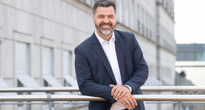 Ein bärtiger Mann in Anzug lehnt sich an ein Geländer.