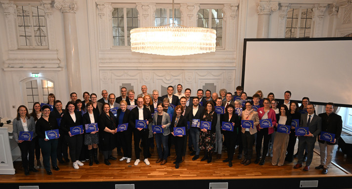 Eine Gruppe an Menschen steht im Halbkreis auf einer Bühne und hält blaue Schilder in der Hand. 