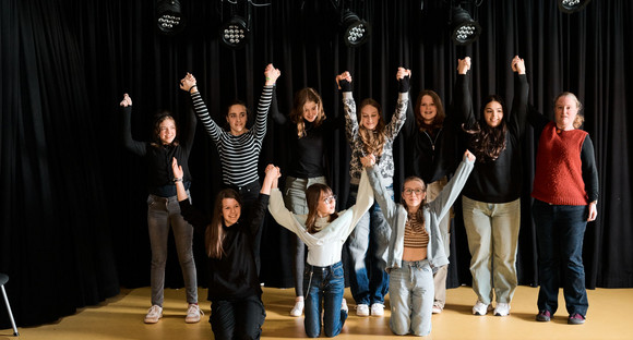 Zehn Schülerinnen im Teenager-Alter stehen in zwei Reihen auf einer Theaterbühne. Sie halten sich an den Händen, die sie gemeinsam in die Höhe strecken und blicken ins Publikum. 