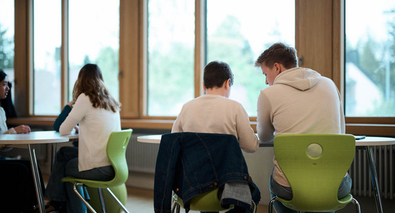 Schüler und Schülerinnen sitzen an Tischen in einem Schulraum.