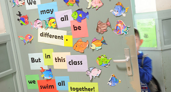 Auf bunten Zetteln, die an einer Tür kleben, steht: We may all be different. But in this class we swim all together! Ausgeschnittene Bilder von bunten Fischen sind ebenfalls an die Tür geklebt.