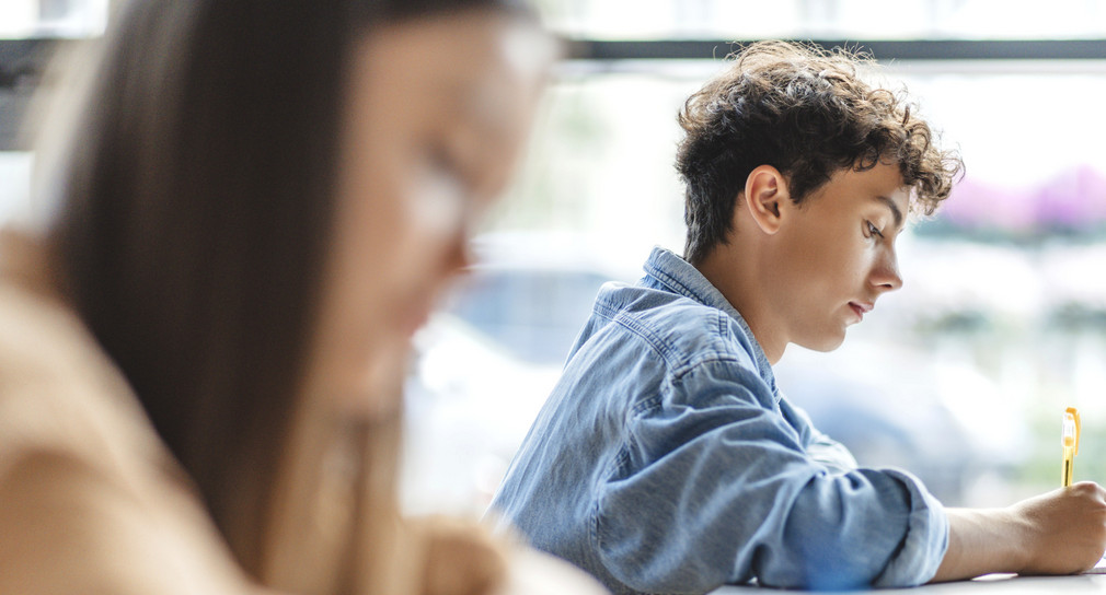 Schülerin und Schüler sitzen nebeneinander und schreiben konzentriert. Der Schüler ist im Focus.