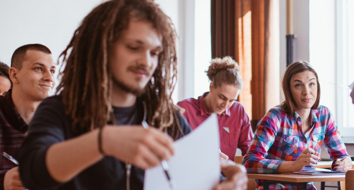 Ältere Schülerinnen und Schüler im Klassenraum. Im Focus ein Schüler, der ein Blatt Papier hochhält