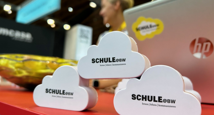 Bild von drei wolkenförmigen Würfeln mit dem Logo der Digitalen Bildungsplattform Schule@BW. Im Hintergrund ist ein Messestand zu sehen.