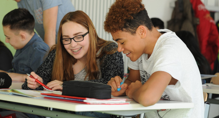 Eine Schülerin und ein Schüler beugen sich zusammen über ein Buch. Sie haben Spaß beim Lernen.