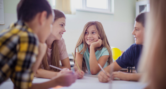 Jugendliche Schülerinnen und Schüler sitzen im Klassenzimmer um einen Tisch und reden und schreiben.