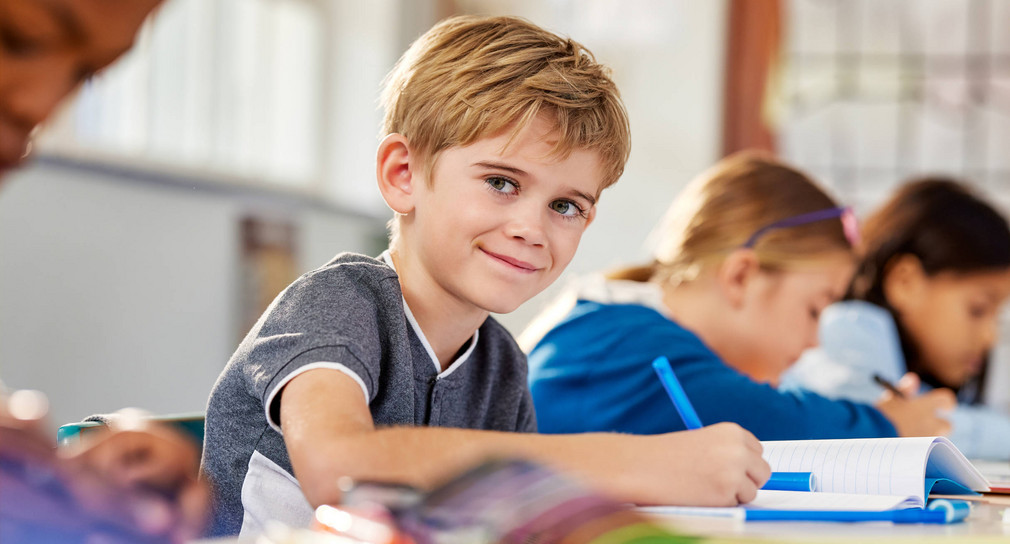 Ein Schüler ein einem Klassenzimmer, der mit einem Stift in ein Heft schreibt. Im Hintergrund weitere Schülerinnen.