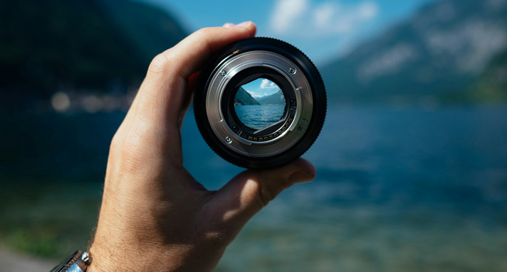 Eine Hand hält eine Objektiv eines Fotoapparates, dahinter sieht man Berge. 