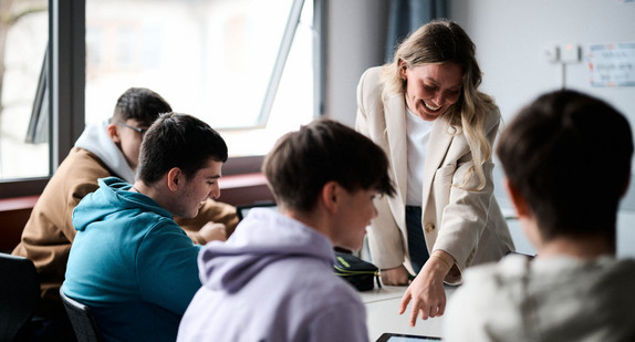 Eine lächelnde Lehrerin zeigt auf ein Tablet, das vor Schülern auf einem Schultisch liegt