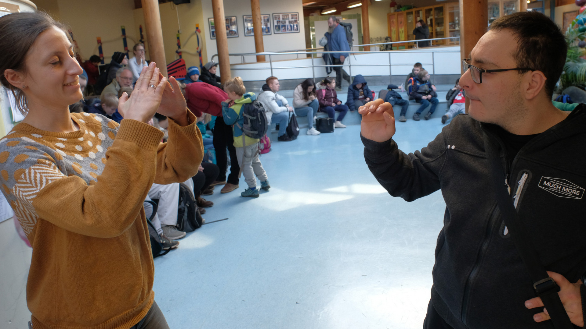 Eine Lehrerin und ein Schüler unterhalten sich in Zeichensprache in einem Schulforum, in dem im Hintergrund  mehrere Schülerinnen und Schüler sitzen