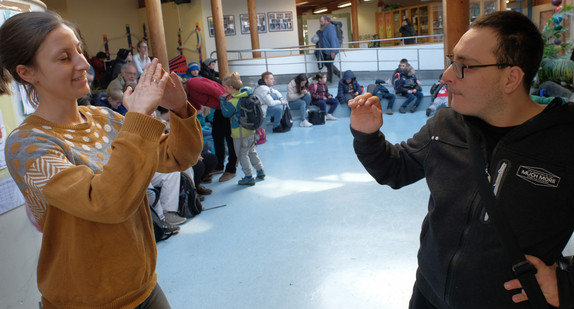 Eine Lehrerin und ein Schüler unterhalten sich in Zeichensprache in einem Schulforum, in dem im Hintergrund  mehrere Schülerinnen und Schüler sitzen