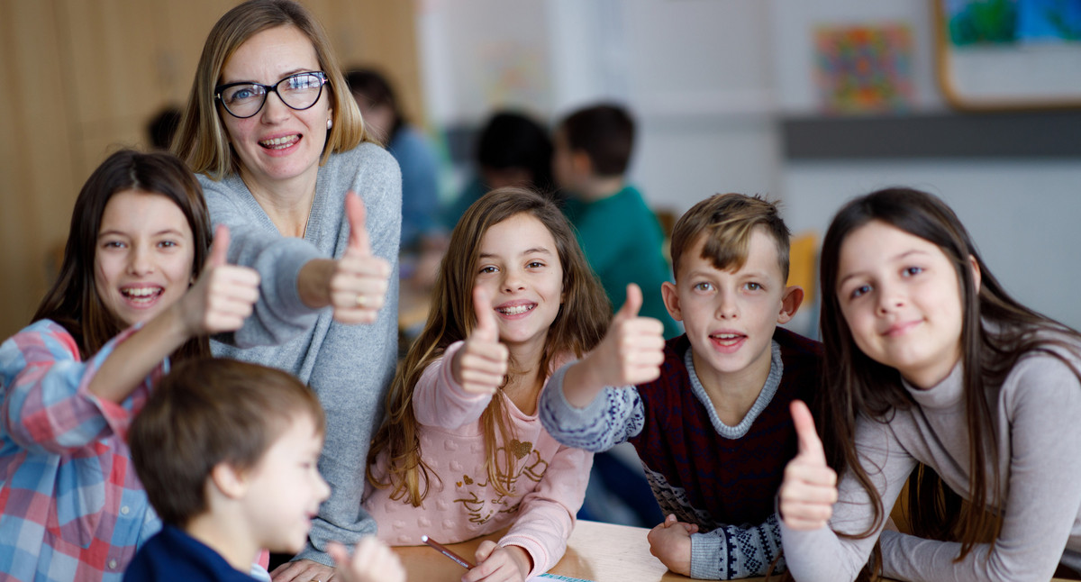 Mehrere Kinder und die Lehrerin sitzen und stehen um einen Tisch und strecken die Daumen nach oben