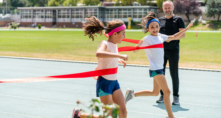 Zwei Mädchen beim Zieleinlauf mit rotem Zielband auf dem Sportplatz, daneben ein Schiedsrichter