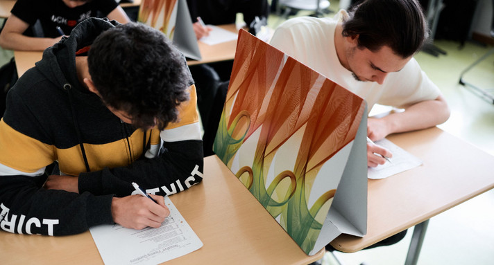 Zwei Schüler sitzen an einem Tisch und schreiben eine Klausur. Zwischen ihnen ist ein Trenner aus Pappe aufgebaut. Beide bearbeiten die Aufgaben auf dem vor ihnen liegenden Papierbogen. 