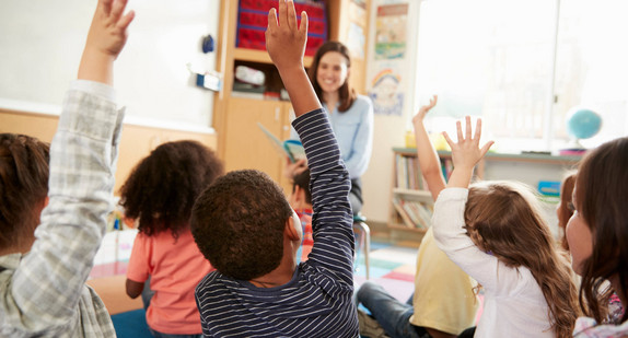 Grundschulkinder sitzen auf dem Boden und heben die Hände. Vor ihnen sitzt eine Lehrerin mit einem Buch