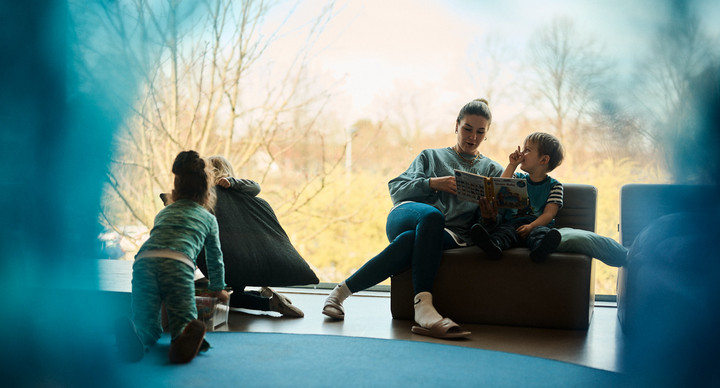 Eine Frau liest einem Jungen aus einem Buch vor, beide sitzen auf einem Sessel vor einem Fenster. Daneben spielen zwei Kinder auf dem Boden. 