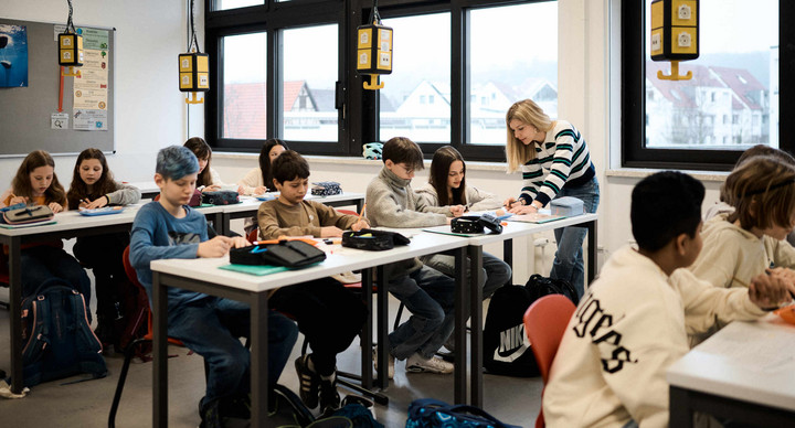 Blick in ein Klassenzimmer, in dem Schülerinnen und Schüler an Tischen sitzen. Eine Lehrerin steht neben einem der Tische.