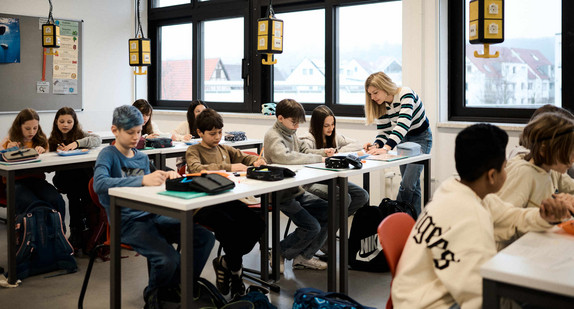 Blick in ein Klassenzimmer, in dem Schülerinnen und Schüler an Tischen sitzen. Eine Lehrerin steht neben einem der Tische.