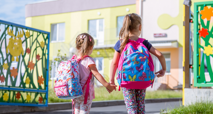 Zwei Mädchen tragen Schulrucksäcke und stehen Hand in Hand vor dem Eingangstor einer Schule.