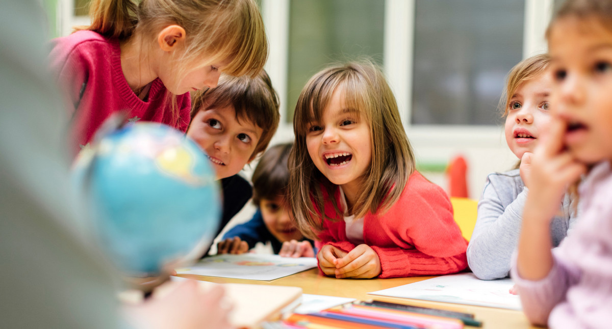 Kinder im Vorschulalter schauen lachend oder gebannt an einen Tisch gelehnt auf eine Lehrerin, die man aber nur im Ansatz von hinten sieht. Ein kleiner Globus steht auf dem Tisch.