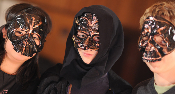 Auf der Schulbühne sind drei Schülerinnen und Schülern mit schwarzen Masken und dunklen, sehr schönen Masken. Der Focus ist auf die maskierten Gesichter gesetzt.