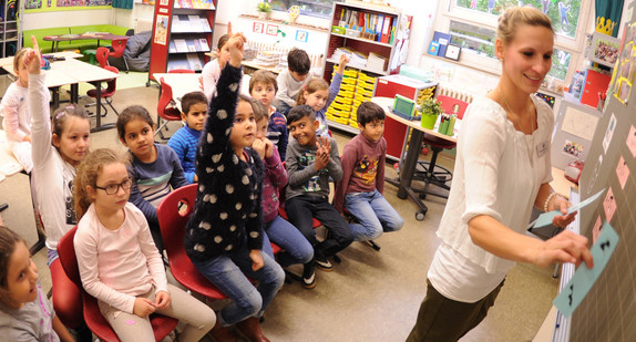Eine eng zusammensitzende Schulklasse ohne Tische schaut auf die Tafel, auf der eine Lehrerin etwas klebt.