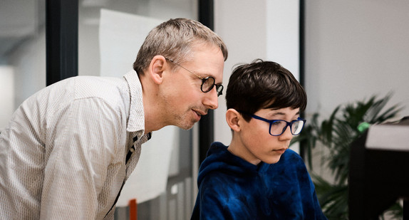 Eine männliche Lehrkraft im mittleren Alter und ein etwa zehnjähriger Schüler blicken gemeinsam konzentriert auf einen Monitor. 