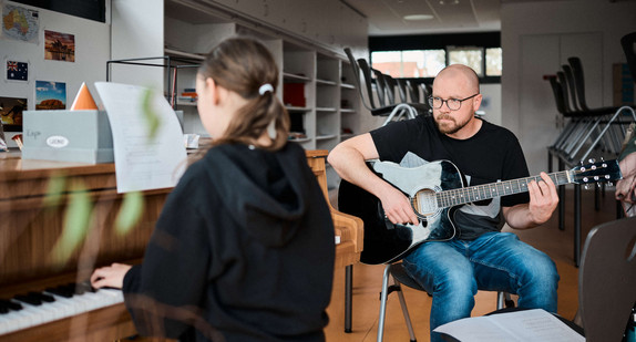 Eine Schülerin sitzt am Klavier, ein Lehrer begleitet sie mit einer Gitarre