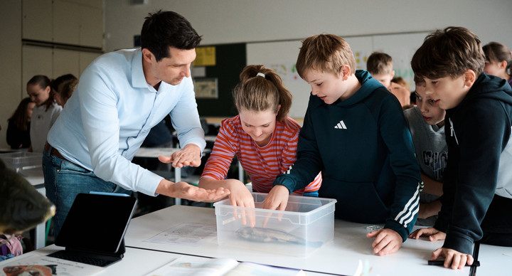 Ein Mann steht neben drei Kindern an einem Tisch, auf dem eine Plastikbox mit einem Fisch drin liegt.   