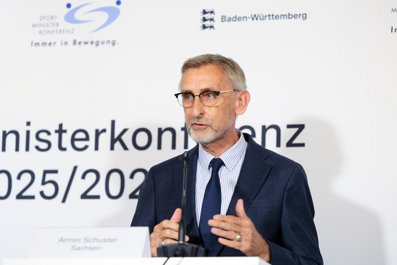 Sportministerkonferenz Sportministerkonferenz 8. und 9. Mai 2025 in Chemnitz Ein Mann mit Anzug und Krawatte steht vor einer weißen Wand, auf der das Logo der Sportministerkonferenz zu sehen ist.