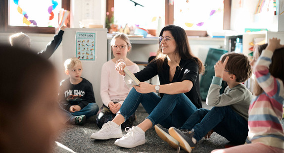 Mehrere Kinder und eine Lehrerin sitzen in einem Schulraum im Kreis auf dem Boden.