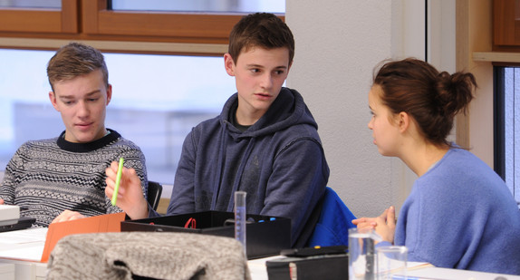 Zwei jugendliche Schüler und eine Schülerin miteinander im Gespräch im Klassenraum