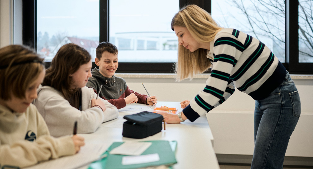 Eine Frau Lehnt sich über einen Tisch, an dem drei Kinder sitzen.