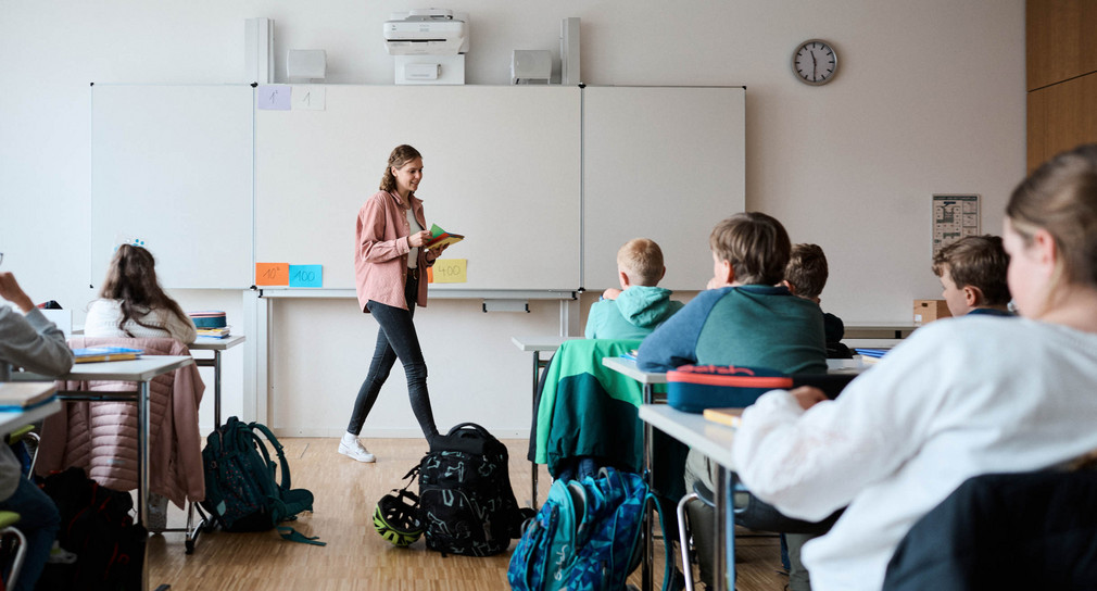 Eine Lehrerin vor einem Whiteboard, ihr gegenüber sitzen Schülerinnen und Schüler an Schultischen