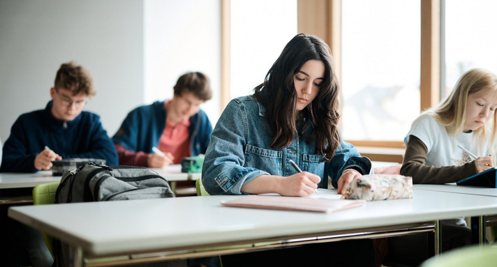 Mädchen sitzt in der Schule an einem Tisch und schreibt. Hinter ihr sind weitere Schülerinnen und Schüler zu sehen.