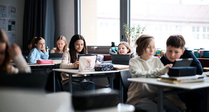 Schülerinnen und Schüler mit sitzen mit Tablets an Tischen in einem Klassenzimmer