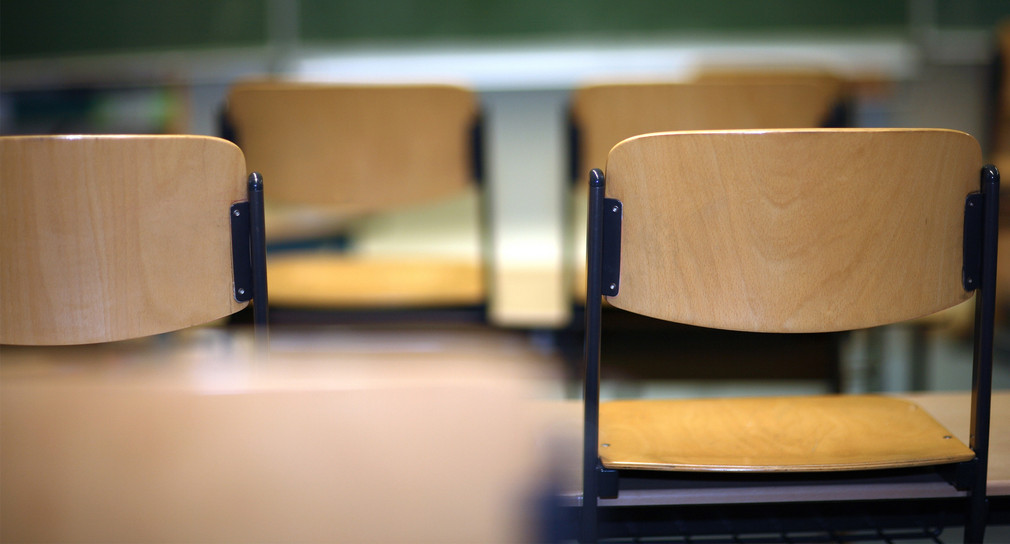 Leere Holzstühle in einem Klassenzimmer, unscharfe grüne Tafel im Hintergrund.