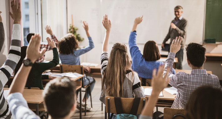 Klasse mit Berufssschülerinnen und -schülern von hinten fotografiert. Die meisten melden sich. Vorne steht ein Lehrer vor der Tafel.