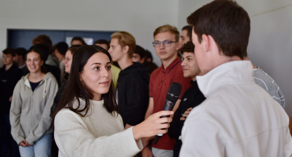 Eine junge Frau hält einem Schüler ein Mikro hin, im Hintergrund stehen weitere Jugendliche