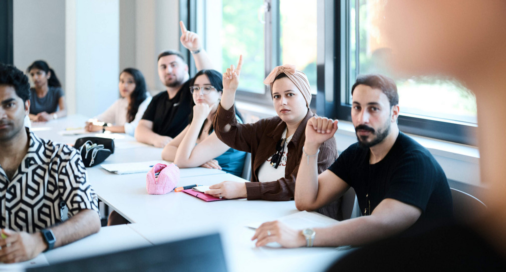 Schülerinnen und Schüler sitzen in einem Schulraum. Einige melden sich.